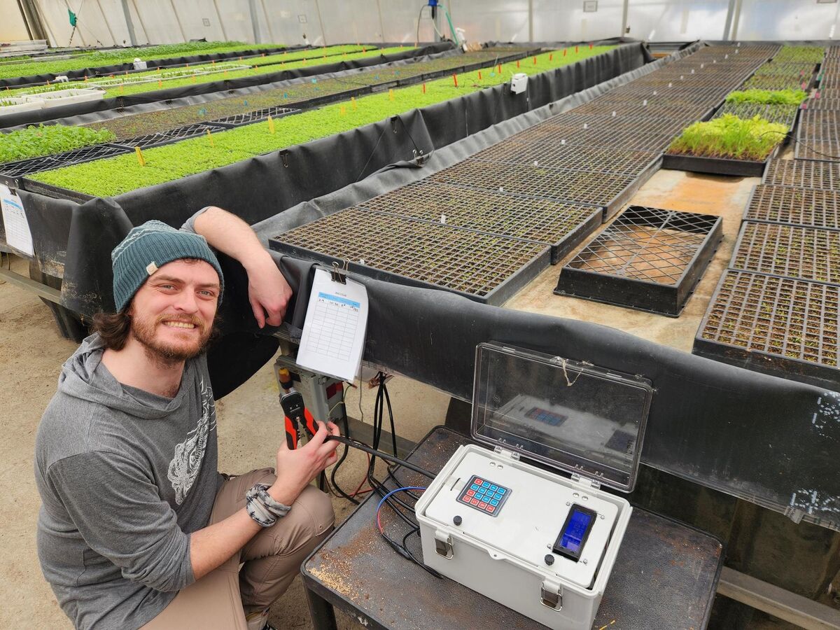 Corey crouching beside a custom watering controller in a greenhouse full of seedling trays